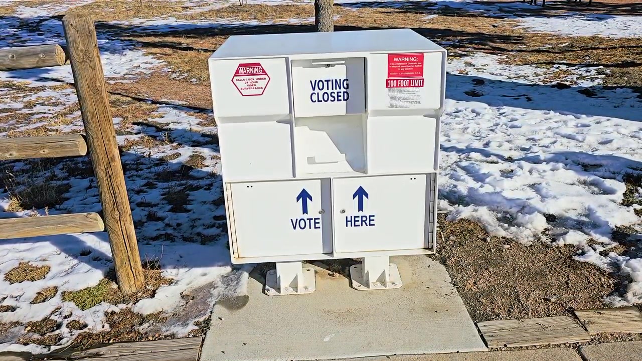 Outdoor mail-in ballot drop box marked Vote Here and Voting Closed, with secure election collection slots, warning signs, directional arrows, snow, and winter sunlight at a polling location.
