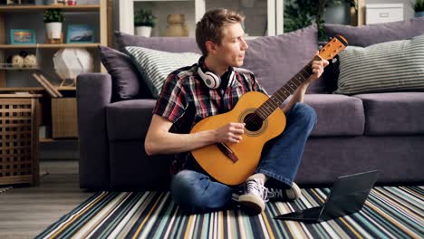 Young man is watching tutorial using laptop and playing the guitar sitting on floor at home