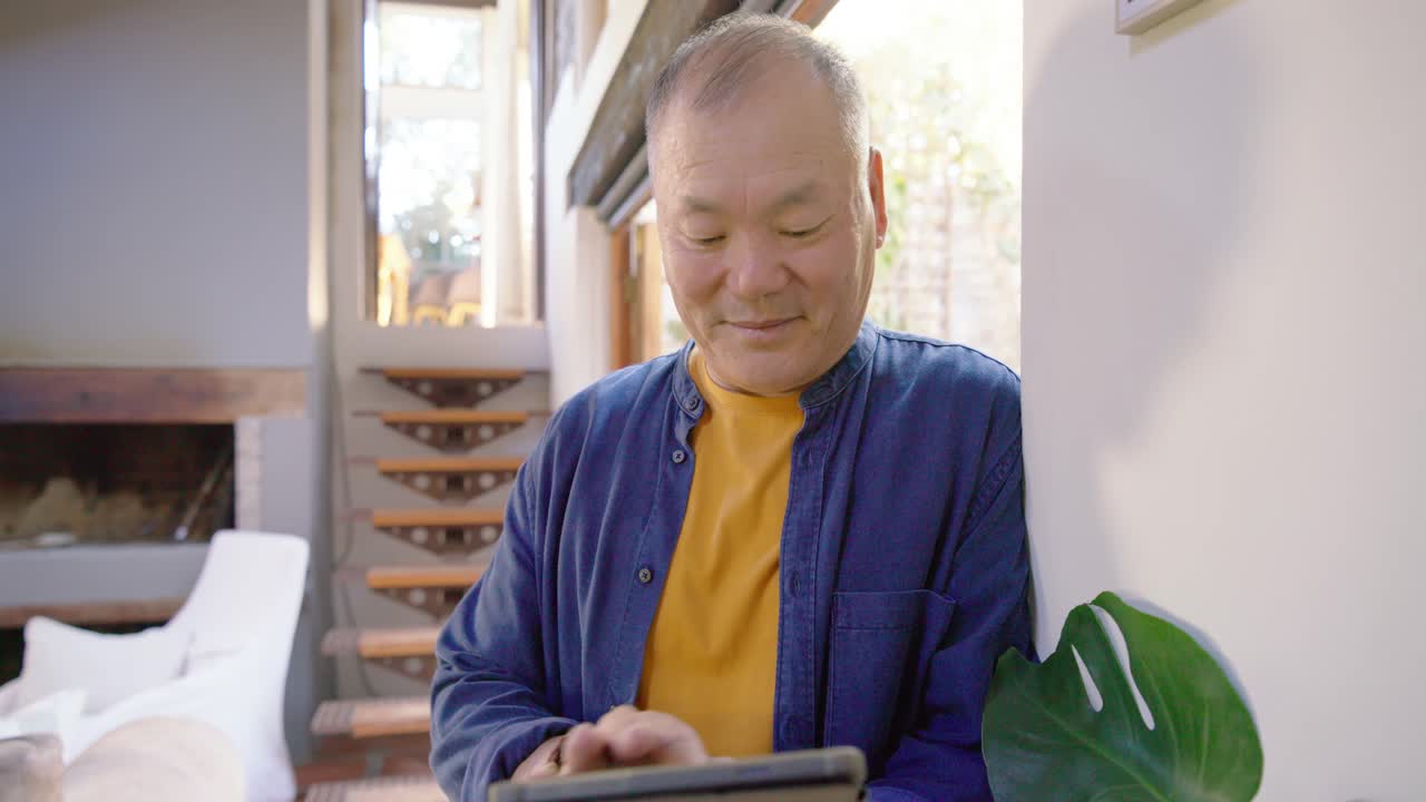 折り戸の近くの壁際に立つシニアのアジア人男性が、タブレットを操作しながら屋外の音に微笑んでいる。