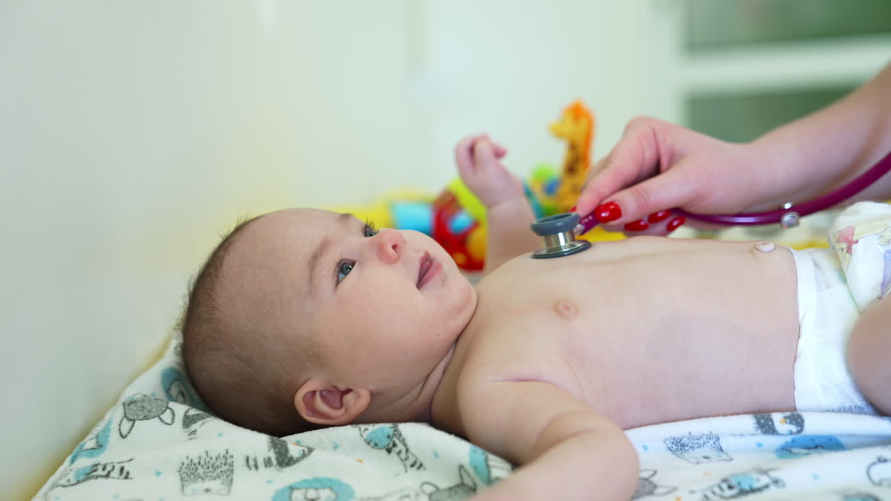 Baby doctor checks little boy's health with stethoscope. Kid looks up cheerfully, waving hands, legs and smiling to the pediatrician. Side view.