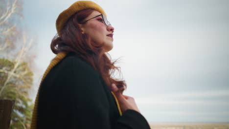 young lady wearing yellow beret and black coat gazes into distance with a serene expression, surrounded by autumn trees with golden leaves against a soft sky