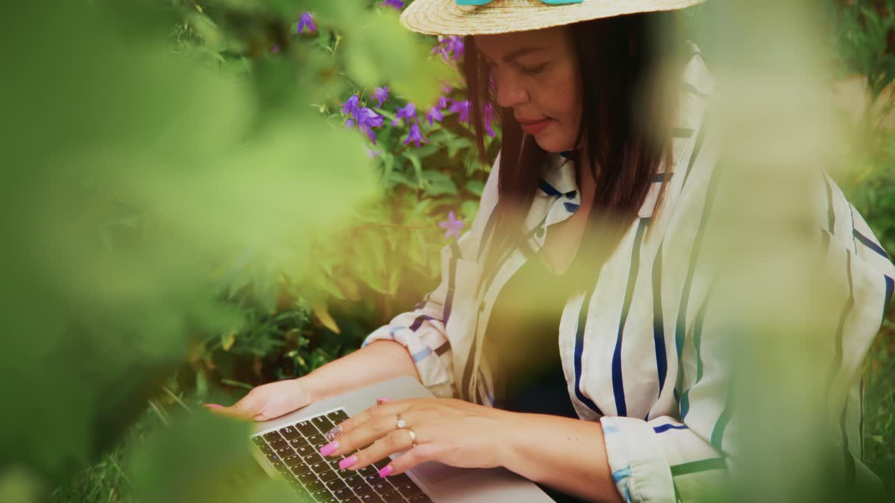 Woman working on laptop in a lush garden surrounded by vibrant flowers and greenery