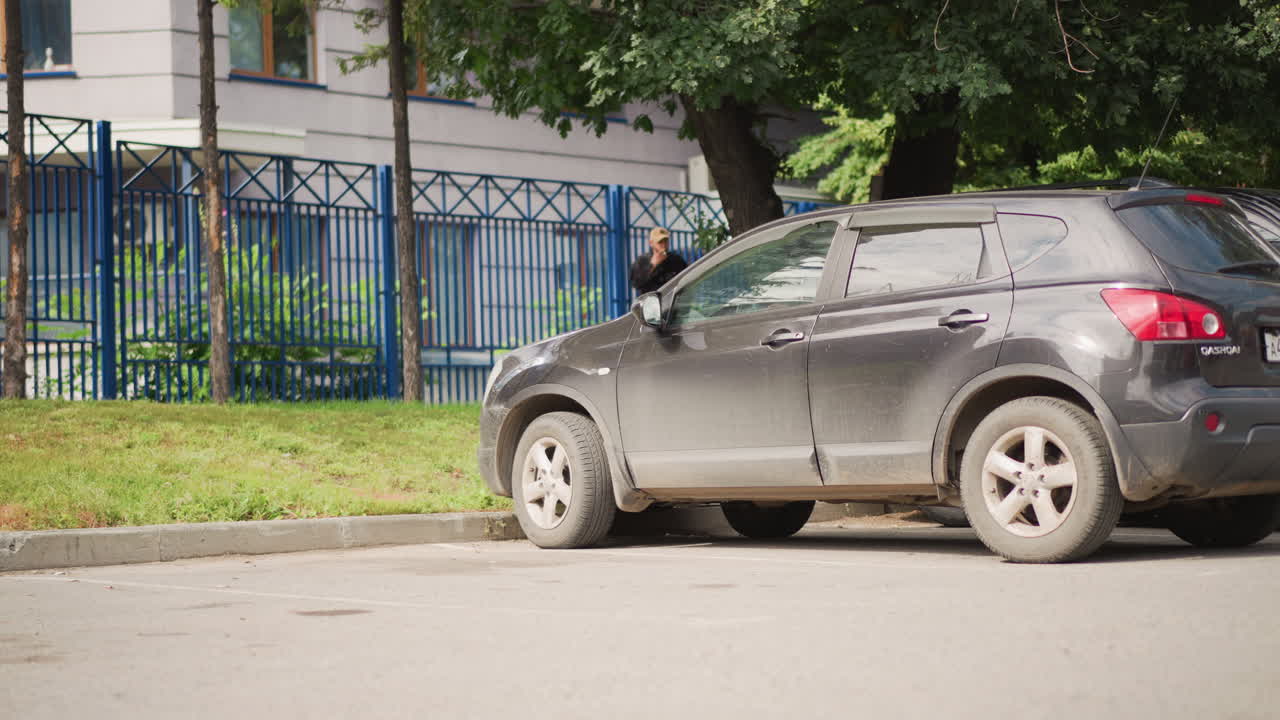 SUV impolverato parcheggiato lungo una recinzione, veicolo fermo al cordolo con ruote schizzate di fango, ombra di albero, strada tranquilla di quartiere, asfalto illuminato dal sole e disegni d'ombra, natura morta urbana adatta per sfondo.