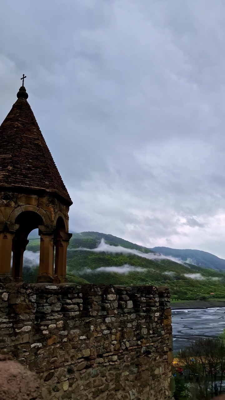 Ancienne tour de la forteresse d'Ananuri surplombant des montagnes brumeuses et un réservoir sous un ciel nuageux spectaculaire.