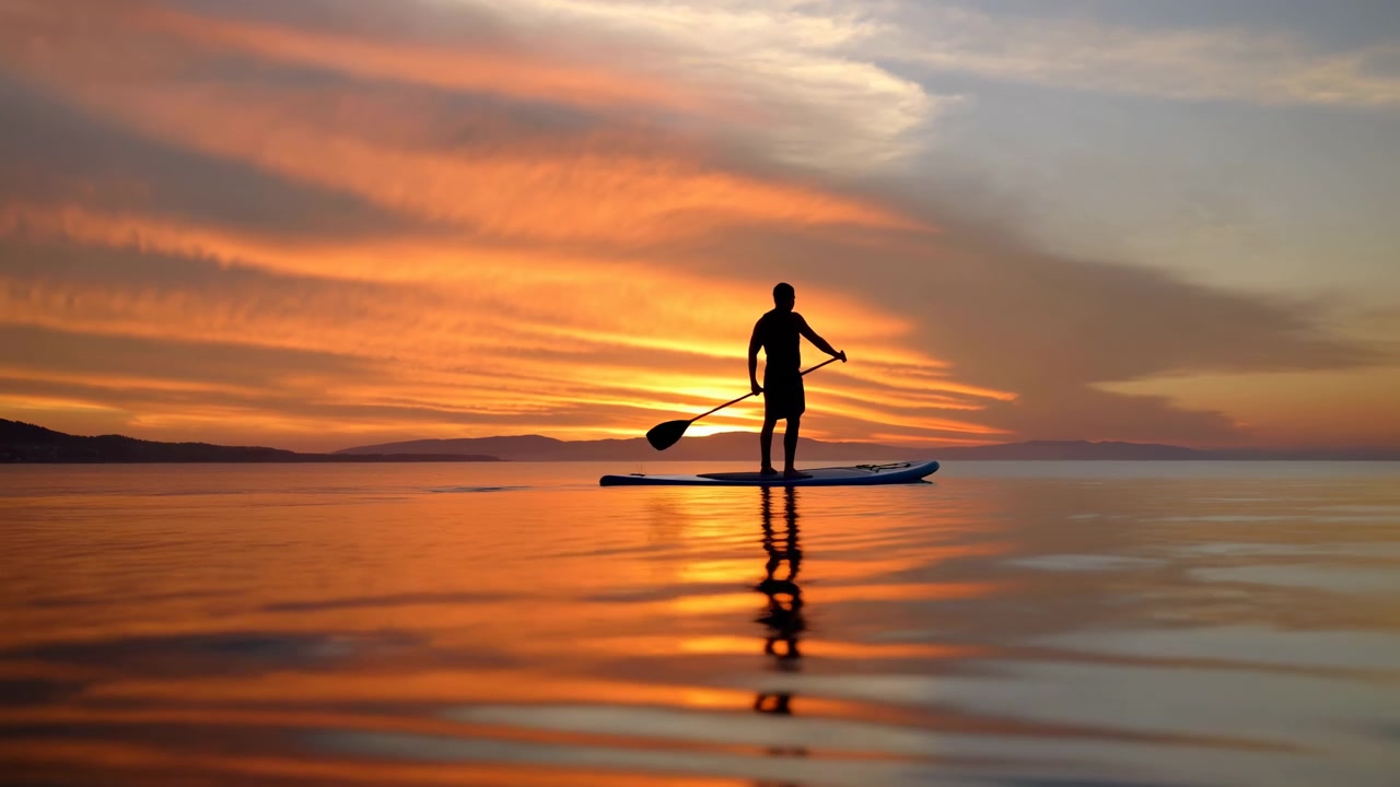 Una persona solitaria in silhouette sta su una tavola da paddleboard vicino al centro di un lago calmo, tenendo una pagaia sotto un drammatico tramonto arancione con luce riflessa