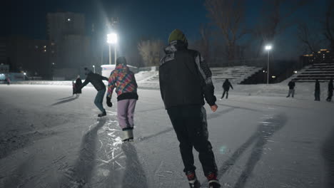 young man skates across icy surface under bright lights with other beginners in background practicing moves near snow covered edges and benches on spacious outdoor ice rink