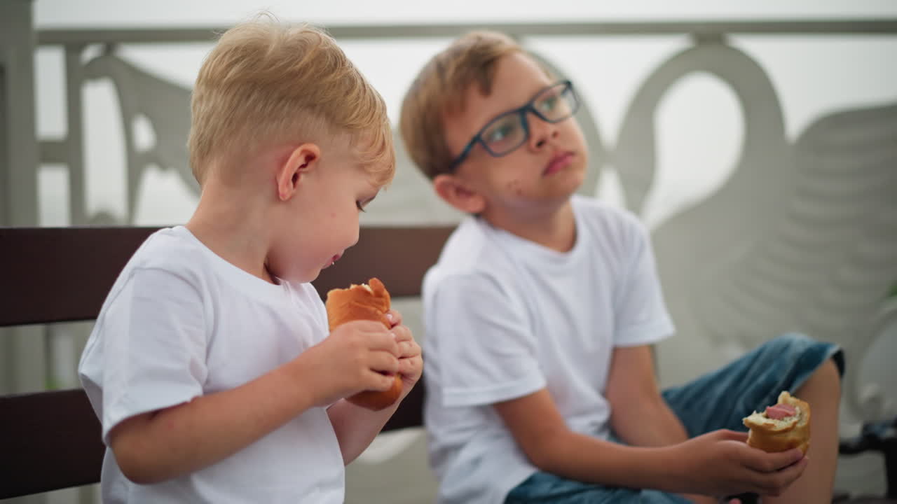 Two happy siblings sit side by side on a bench, joyfully eating sausage rolls, the older brother raises his leg slightly with a serious face while the younger sibling lifts himself in excitement