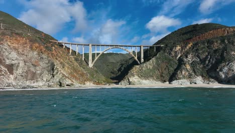 toma cinematográfica del puente bixby contra la cordillera verde mientras las olas chocan contra la orilla, big sur california