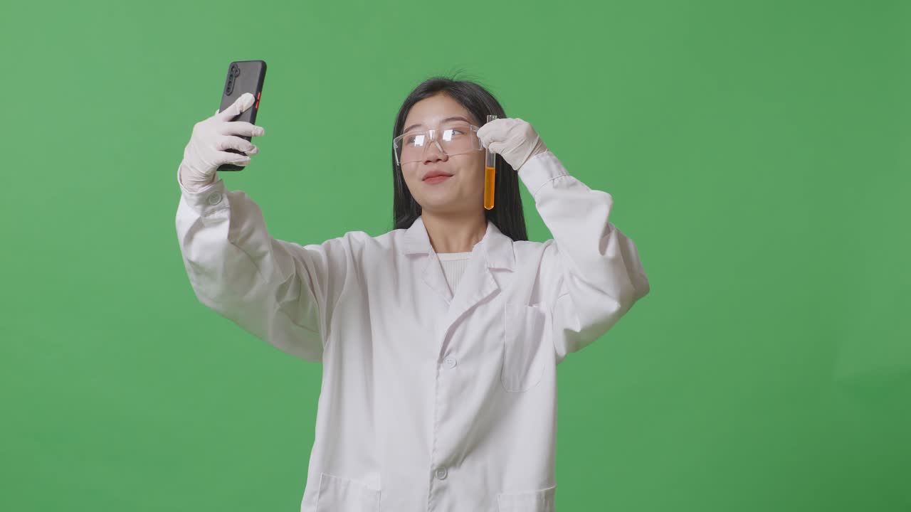 아시아 여성 과학자가 오렌지색 액체의 테스트 튜브를 들고 실험실의 녹색 화면 배경에 서서 스마트폰에서 비디오 통화를 하고 있습니다.