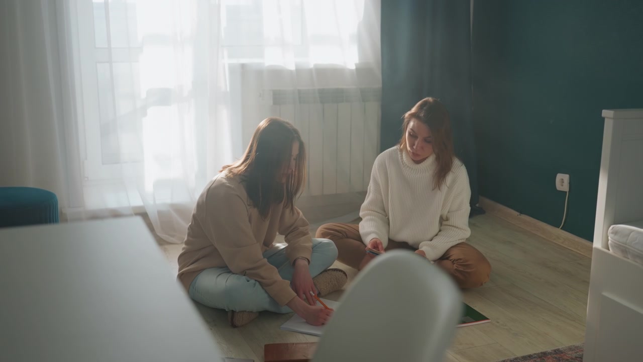 To kvinder studerer på gulvet i solbeskinnet stue med bærbar computer, notesbøger, kuglepenne og grammatikbog, venlig vejleder giver feedback, afslappede trøjer, varmt naturligt lys, rolig og støttende atmosfære