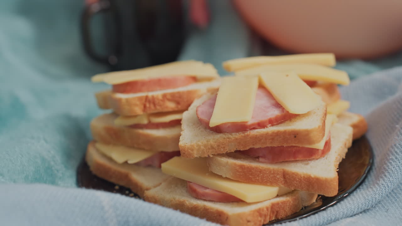 Close up of multiple bread slices layered with cheese and sausage arranged neatly on dark plate placed over soft colorful fabric, hand in background reaching towards food