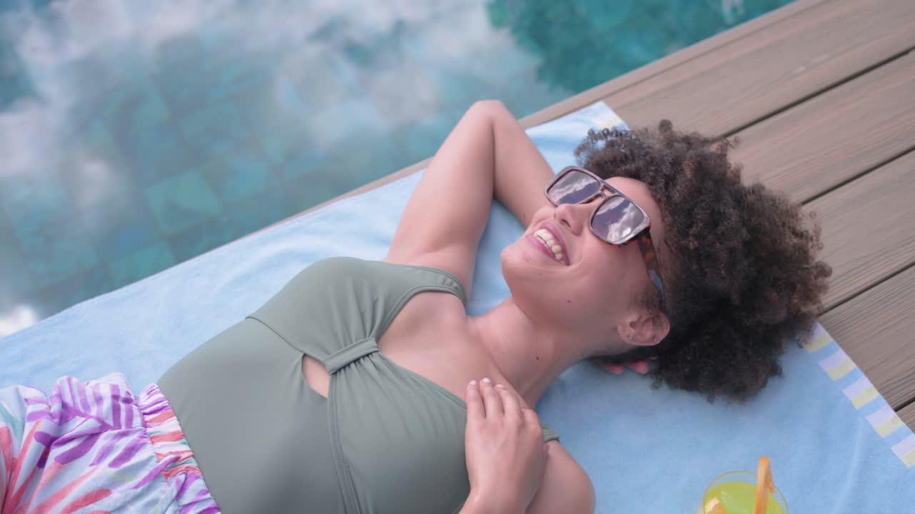 Mulher africana adulta tomando sol, virando-se, rindo, movendo o braço e ajustando a alça do traje de banho em uma toalha à beira da piscina.