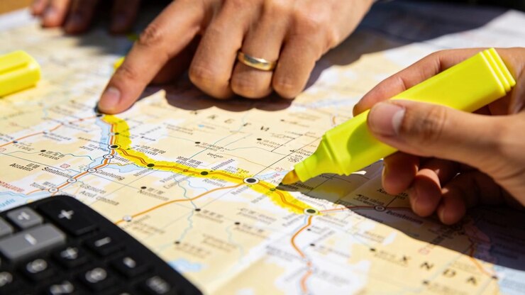 Drawing hands tracing route planning trip at table as yellow marker touching folded map, calculator