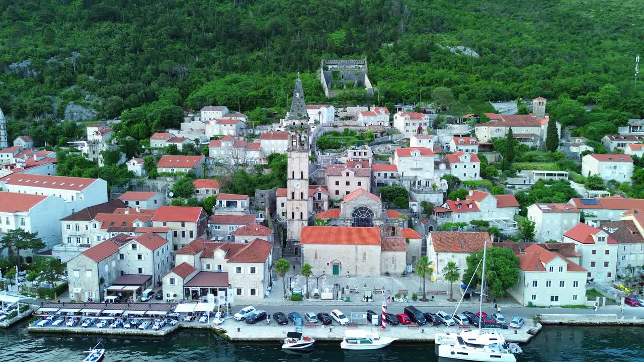 Perast town on bay of Kotor, Montenegro. waterfront restaurant patios with mountain forest in background, Saint Nicholas catholic church bell tower in center, Aerial pullback shot