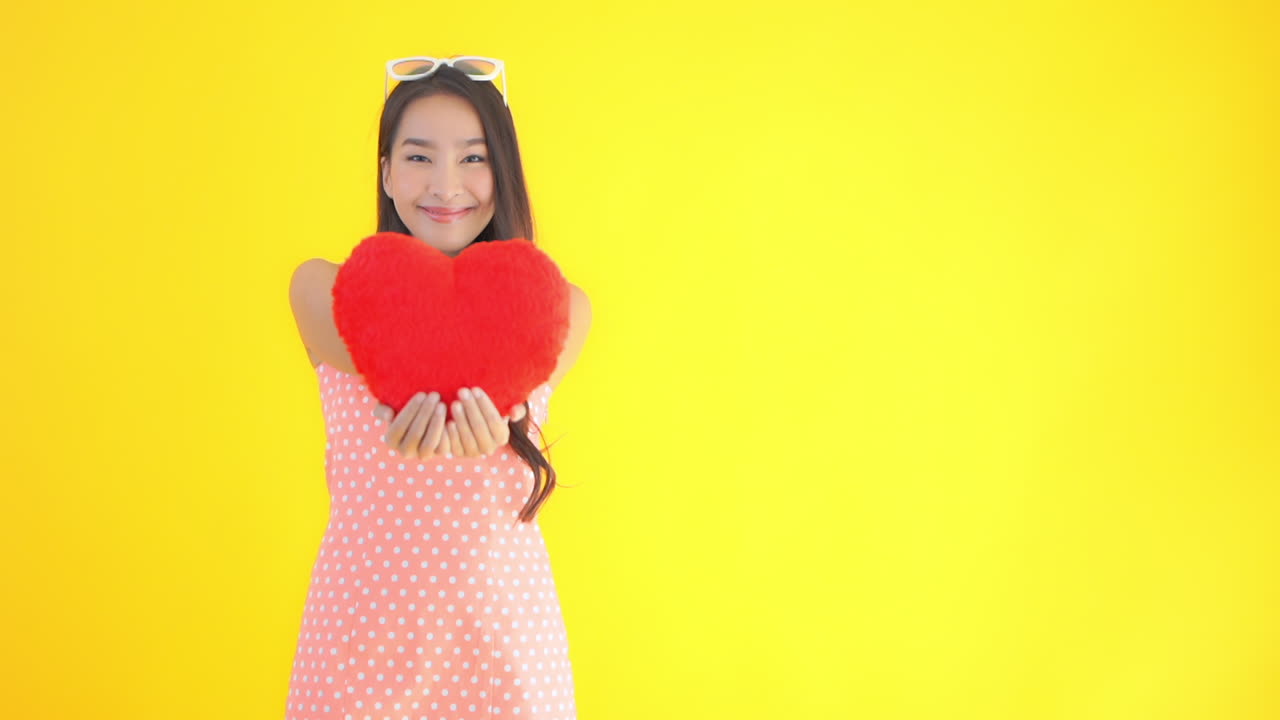 Asian woman in summer pink polka dot dress showing fluffy red heart cushion smiling on a yellow studio background
