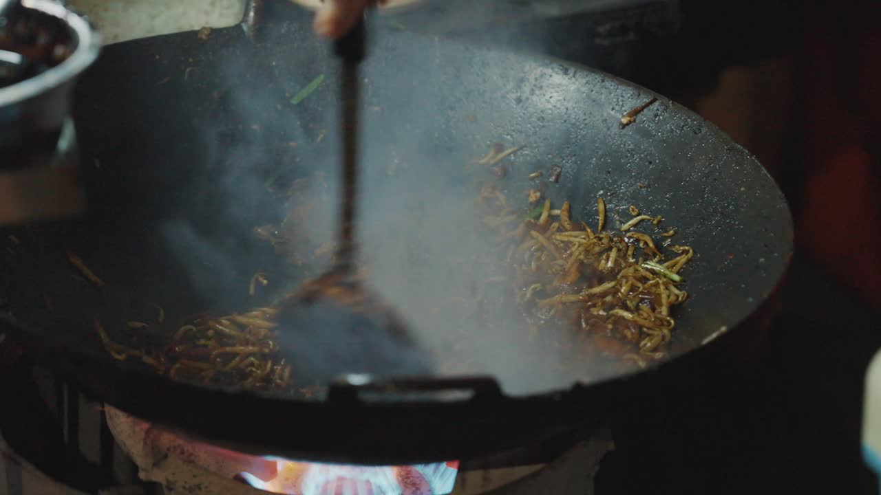 Una ripresa d'azione ravvicinata di un grande wok di ferro stagionato, pieno di vapore e fumo che salgono, mentre un venditore di cibo da strada salta in padella i noodles Char Koay Teow in un affollato mercato notturno.