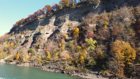 Crystal-clear-blue-river-meanders-beside-a-majestic-mountain-cliff-adorned-in-autumn-foliage,-creating-a-picturesque-and-serene-landscape