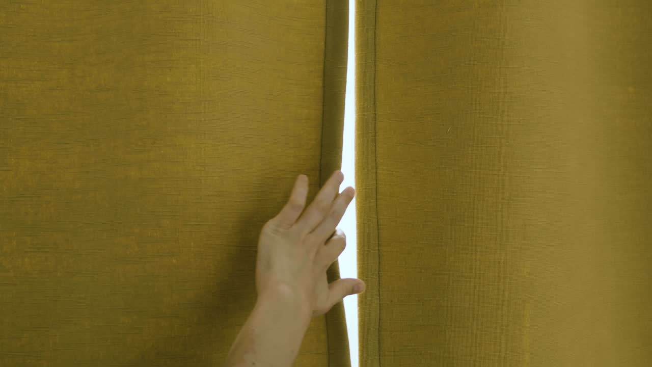 una mano abre lentamente una cortina ocre que cubre una ventana, por lo que entra una luz filtrada y delicada