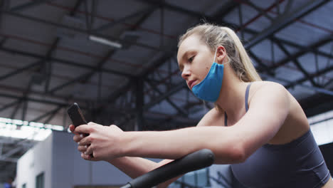 Kaukasische-Frau-Mit-Abgesenkter-Gesichtsmaske-Benutzt-Smartphone-Im-Fitnessstudio