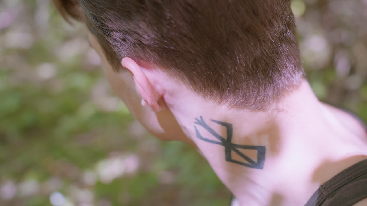 Side view of adventurer loosening headband while glancing in two directions to decide which path to follow, revealing bold tattoo on neck, focused and prepared for upcoming challenges in nature