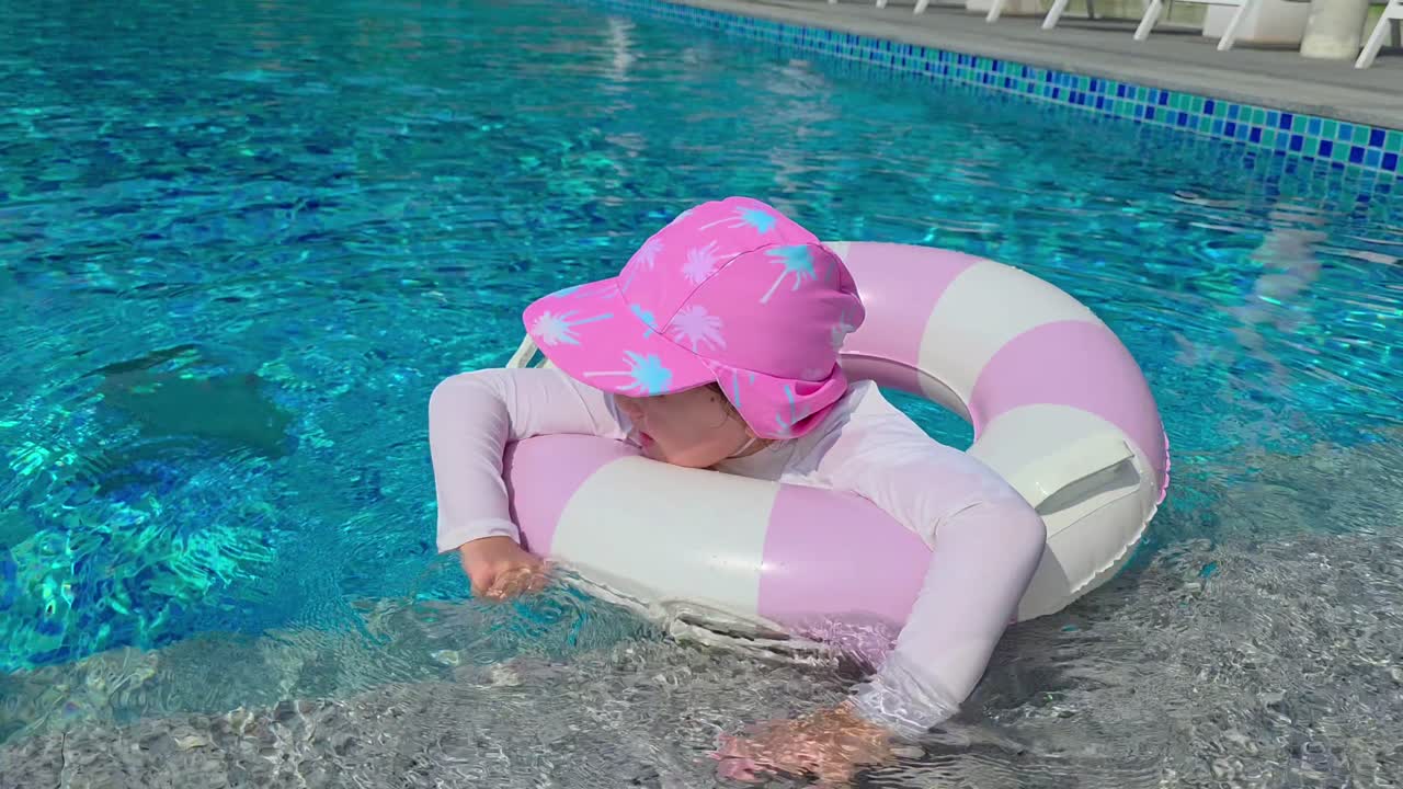 A cheerful Korean-Ukrainian 5 year old girl in a pink sun hat waves, makes a funny face, and flashes a V sign while floating in a blue swimming pool with a pink and white swim ring