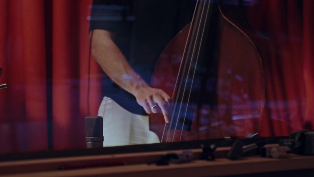 Close-up of a musician plucking a double bass during a studio recording session, with microphone visible and warm red acoustic panels in the background.