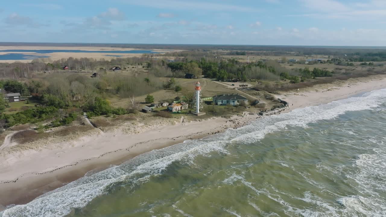 vista aérea del faro de color blanco, costa del mar báltico, letonia, playa de arena blanca, grandes olas que se estrellan, día soleado con nubes, toma de órbita amplia de drones