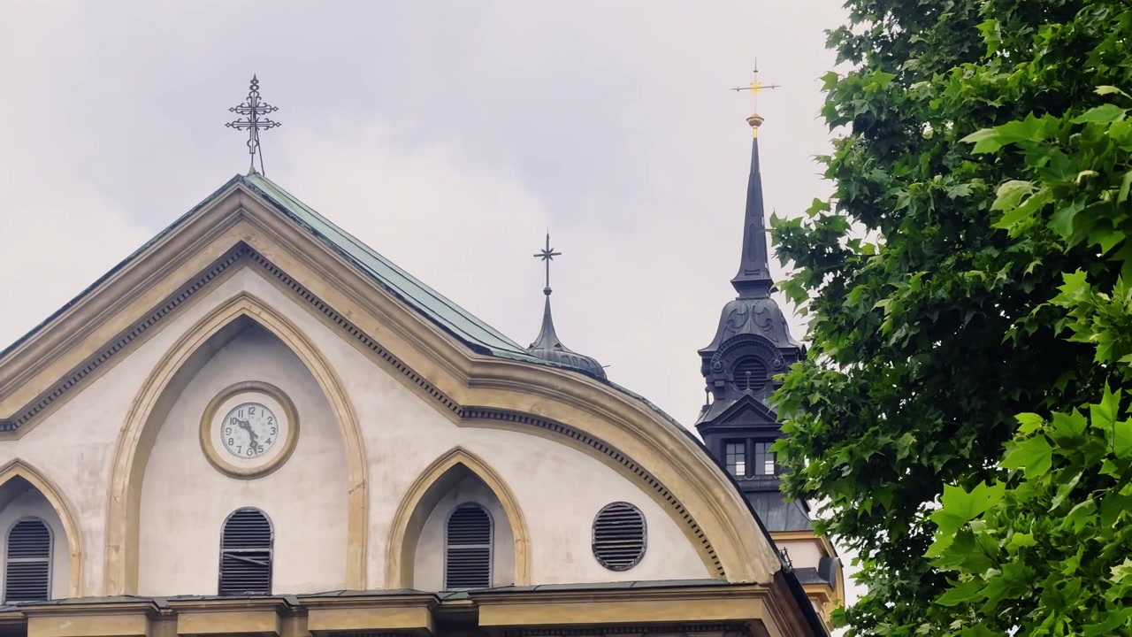 Vista teleobjetiva aproximada da Igreja Ursulina da Santíssima Trindade em Ljubljana, mostrando seu frontão curvo com relógio e pináculos escuros emoldurados por folhas verdes sob um céu nublado