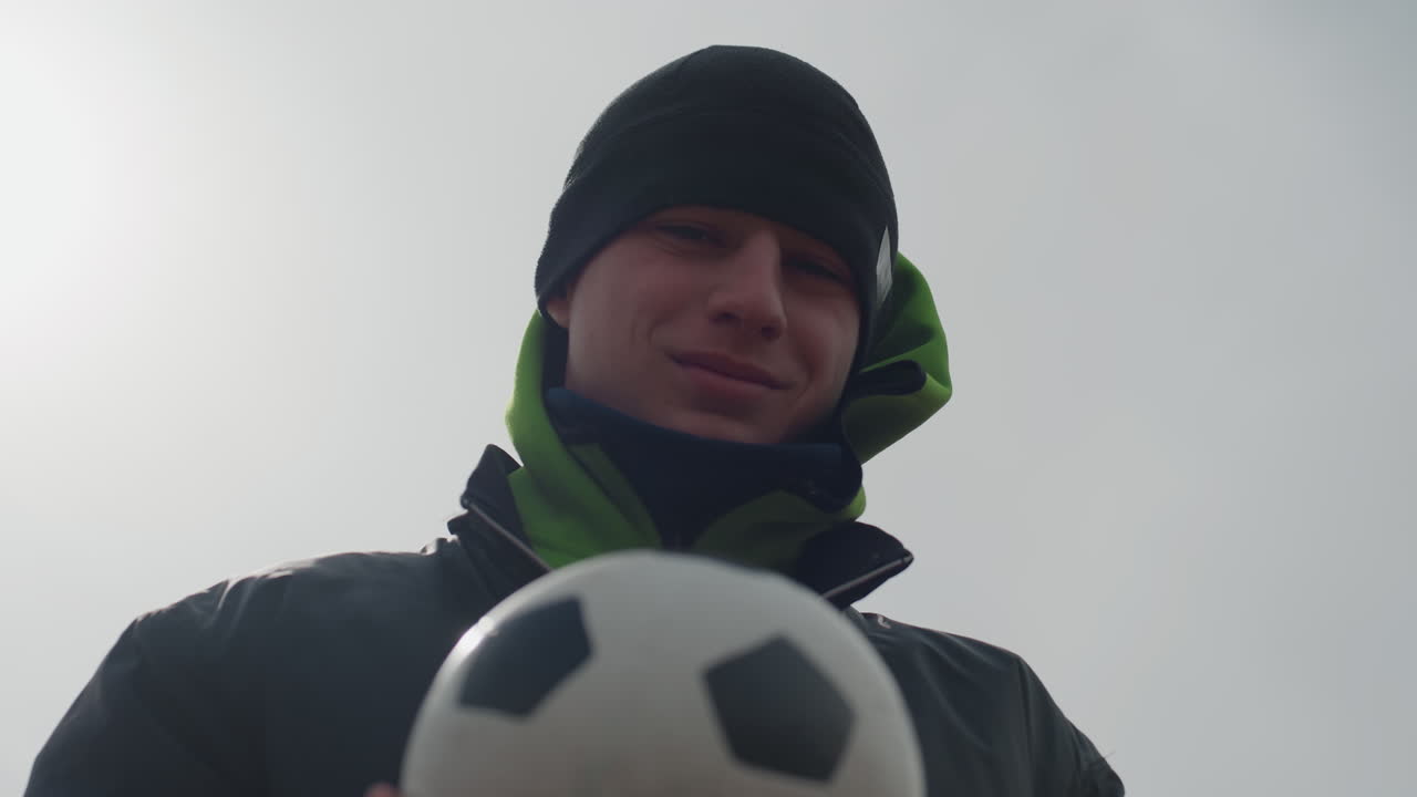 도심 친화적인 축구, 쾌활한 남성이 행복하게 축구공을 보여주는 밝은 도심 풍경, 햇볕이 잘 드는 도시 배경과 편안한 분위기 속에서 축구공을 제시하는 웃는 개인의 클로즈업