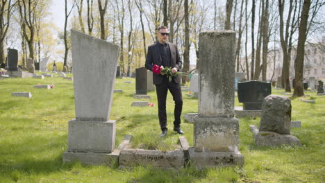 sad man in black suit holding red roses and walking in a graveyard, then taking sunglasses off and looking at tombstones sorrowfully