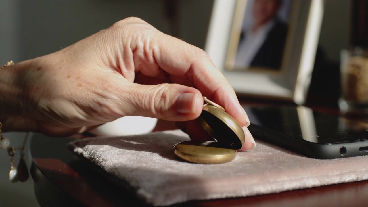 Touching senior hand with pearls opening brass locket on bedside table to view interior near phone