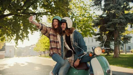 Happy-brunette-girl-in-a-checkered-shirt-and-jeans-takes-a-selfie-using-her-phone-with-her-boyfriend-who-is-sitting-on-a-moped-in-a-sunny-summer-city