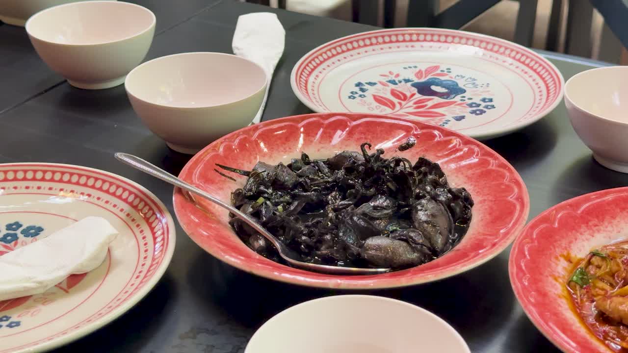 A vibrant Thai dining table displays a red plate of squid in black ink, surrounded by bowls, plates, and utensils under soft indoor lighting with a steady camera angle