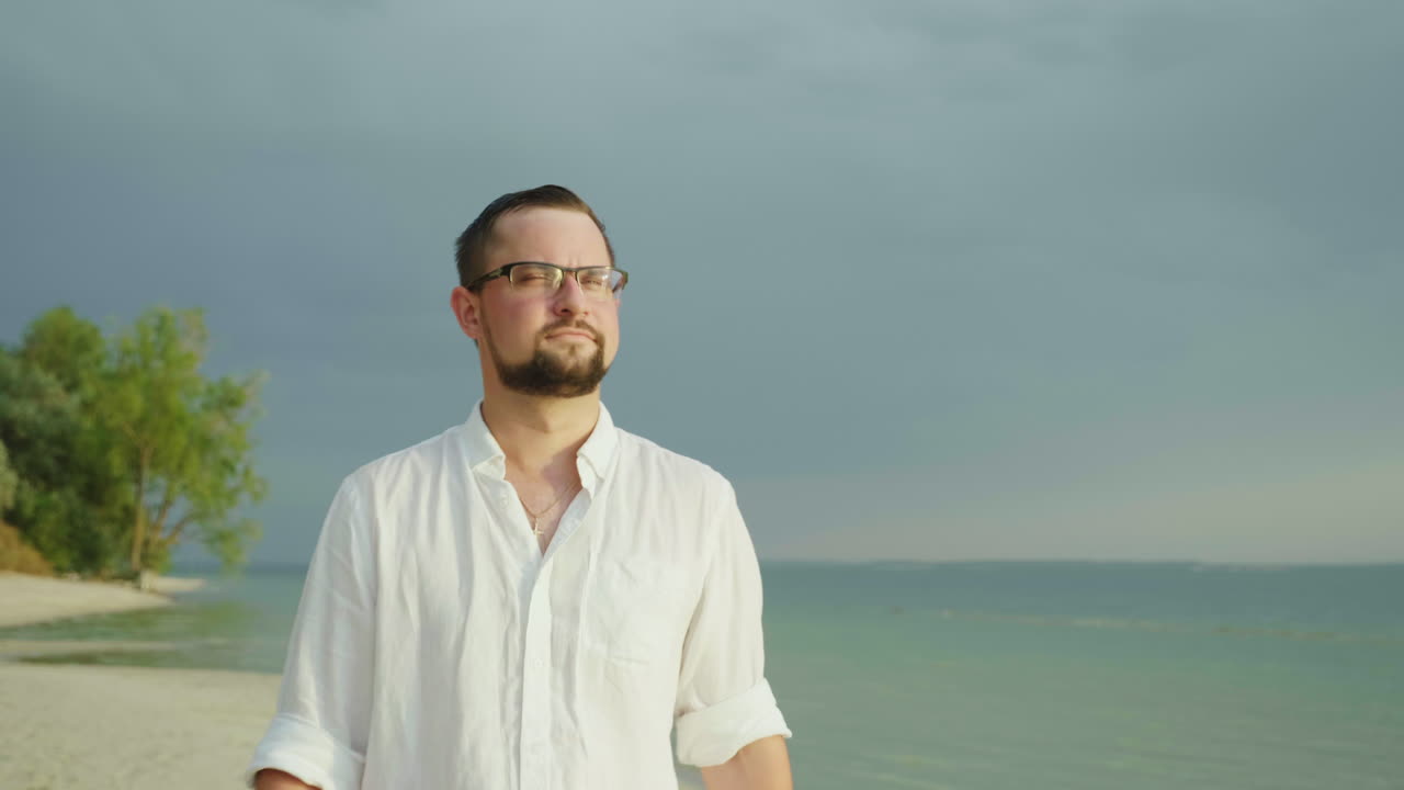 쉬운 셔츠를 입은 한 청년이 열대 리조트 4k 슬로우 모션 비디오에서 해변 휴가를 따라 걷는다