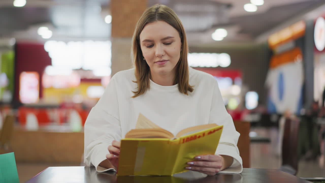 joven leyendo un libro amarillo brillante con una sonrisa sutil en un restaurante moderno, rodeado de un fondo borroso vibrante, con una vista borrosa de otros compradores