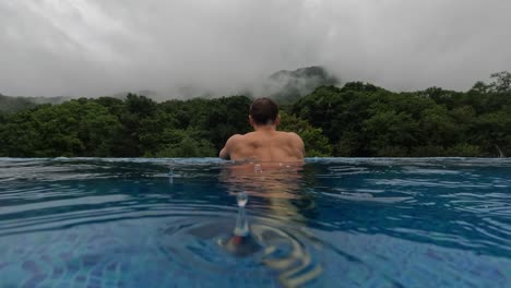 a muscular man walks inside infinity pool with lush mountain view on rainy day