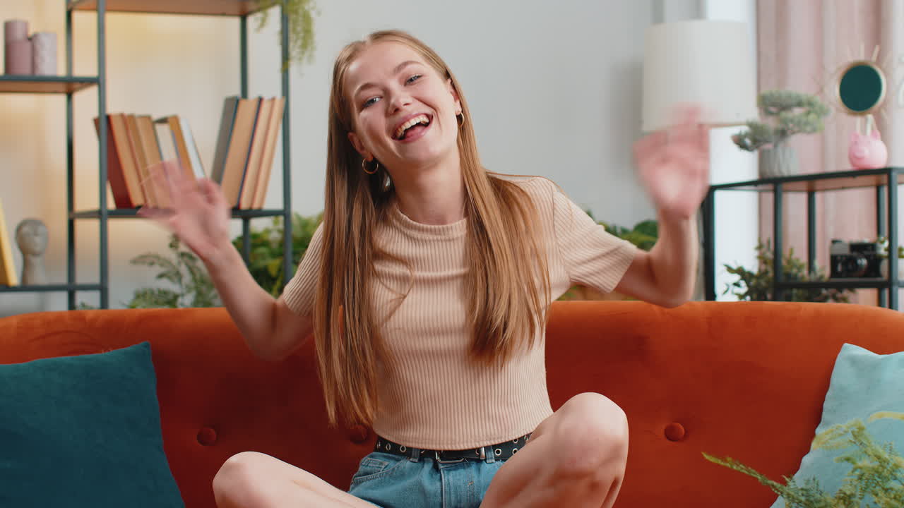 Pretty woman smiling friendly at camera, waving hands gesturing hello, hi, greeting at home on sofa
