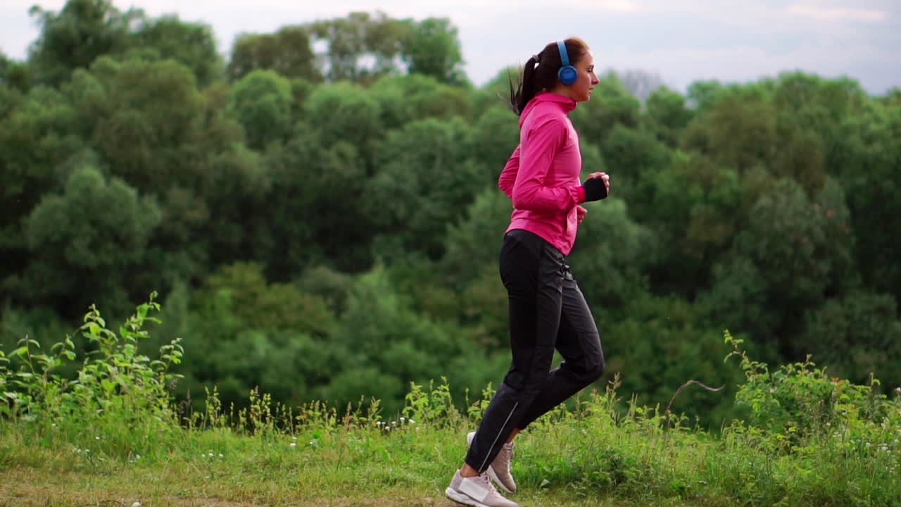 A girl in a pink jacket and black pants runs near the river in headphones preparing for the marathon
