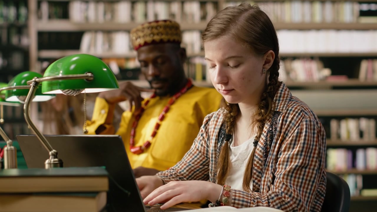 Woman learner studying at shared library table with laptop and open book