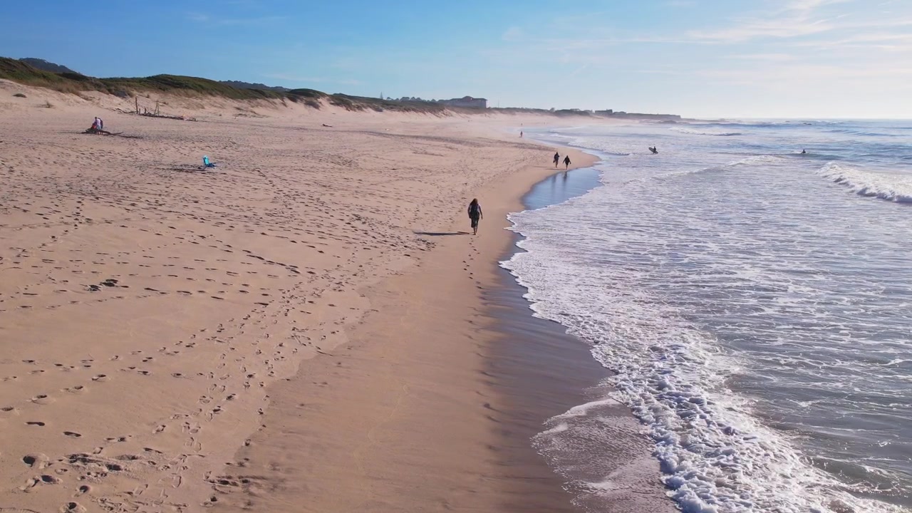 Luftfoto i 4K sakte film som fanger en enslig person som går nær havet, langs en sandstrand med milde bølger, en vid horisont og en fredelig atmosfære.
