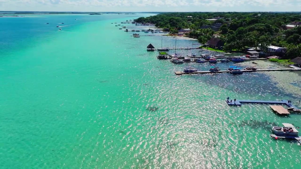 Luftfoto af Bacalar-lagunens kystlinje, Quintana Roo, Mexico, med palapaer, både og moler ved siden af det klare turkise lavvandede område.