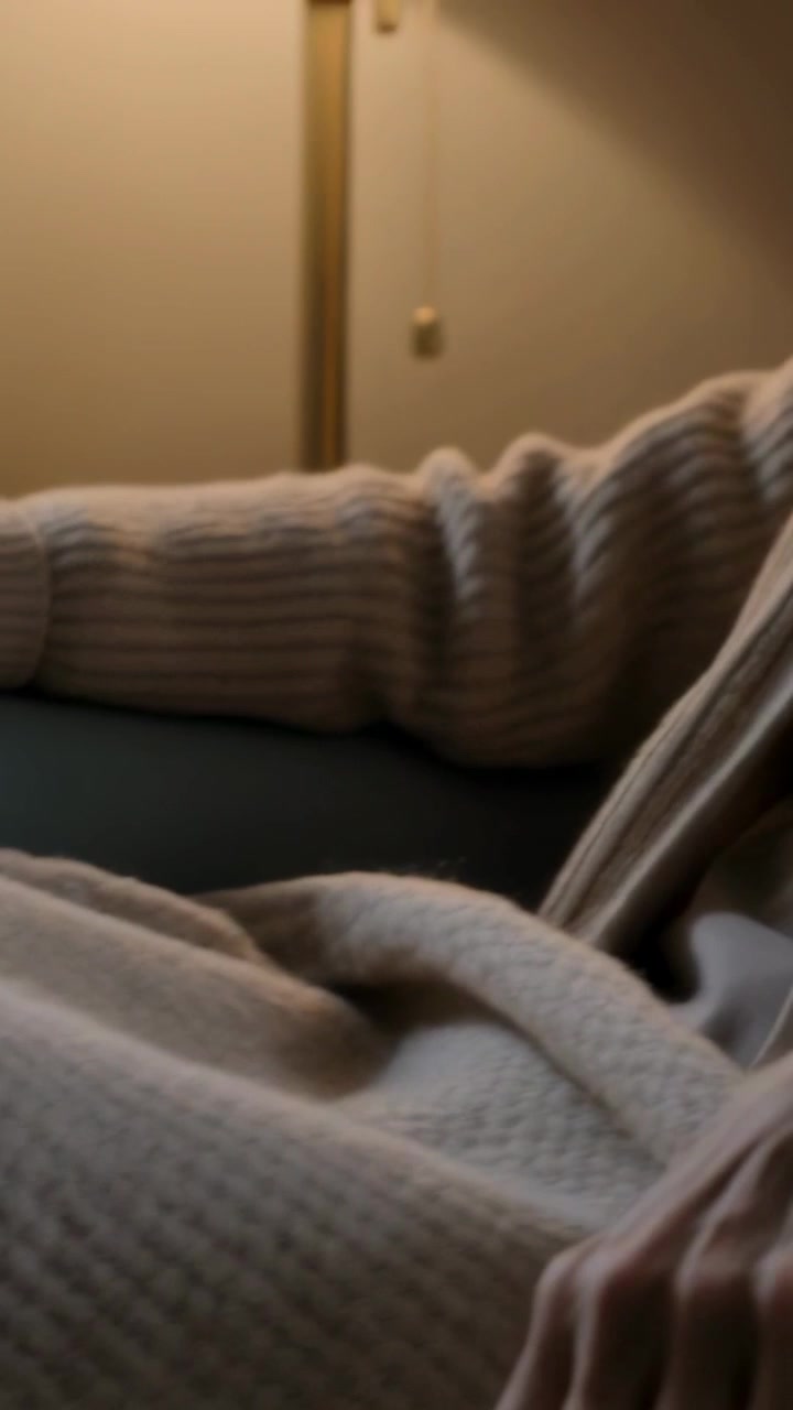 Video verticale: Afferrando il bordo di una coperta, una donna con un maglione a coste si sistema in grembo sulla sedia per riposare accanto a una lampada.