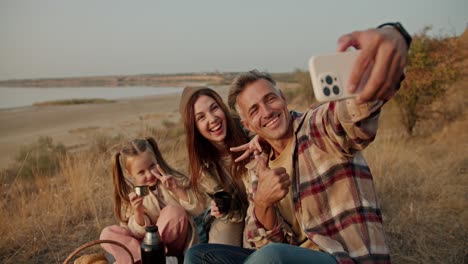 A-happy-brunette-man-with-gray-hair-in-a-plaid-shirt-takes-a-selfie-using-a-phone-of-himself-with-his-wife-and-little-daughter-during-his-picnic-outside-the-city-in-the-summer