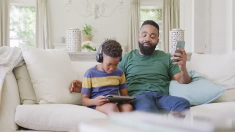happy african american father and son sitting on sofa, using smartphone and tablet, in slow motion