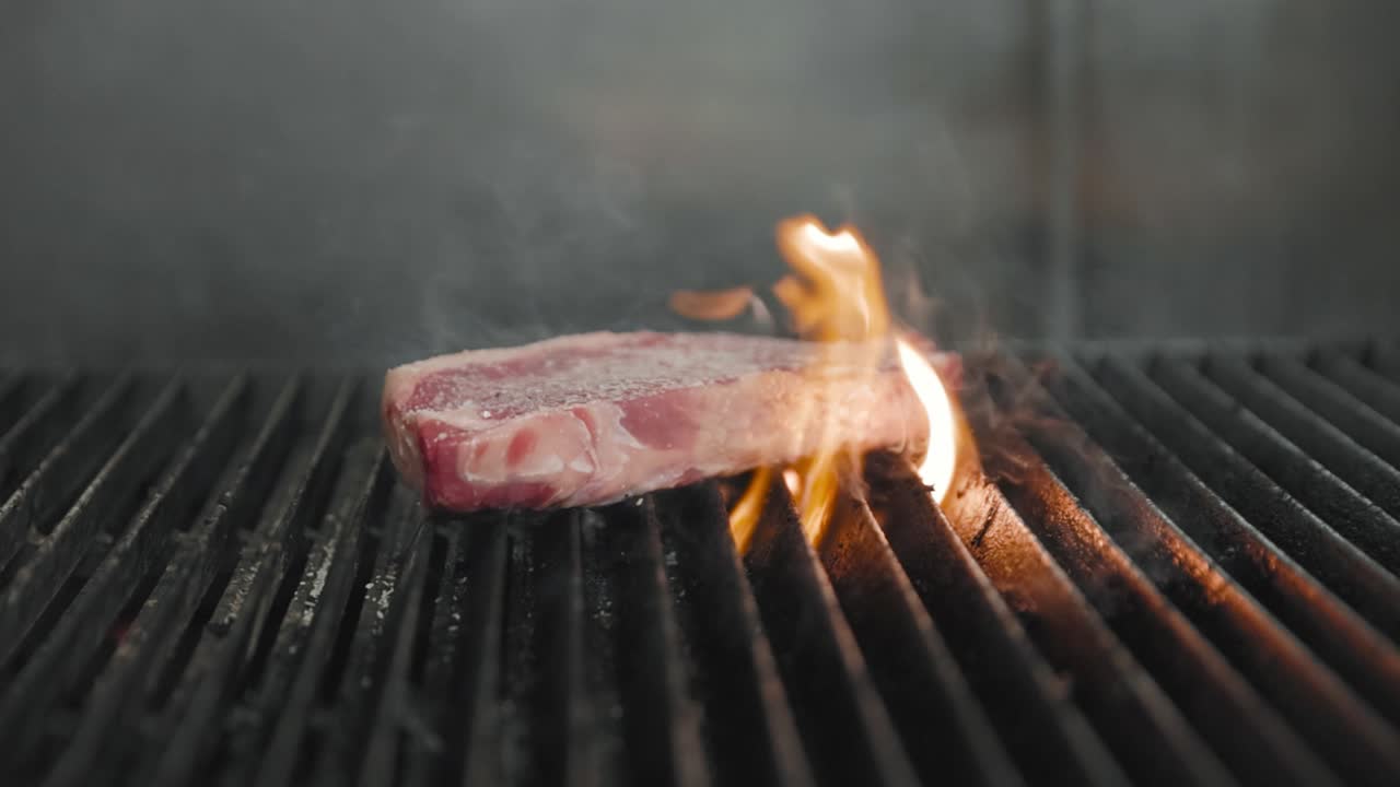 A raw steak sizzles on a grill as open flames rise beneath it, capturing the intensity and flavor or live-fire cooking