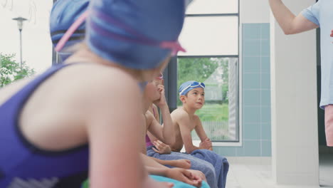 Male-Coach-Giving-Children-In-Swimming-Class-Briefing-As-They-Sit-On-Edge-Of-Indoor-Pool