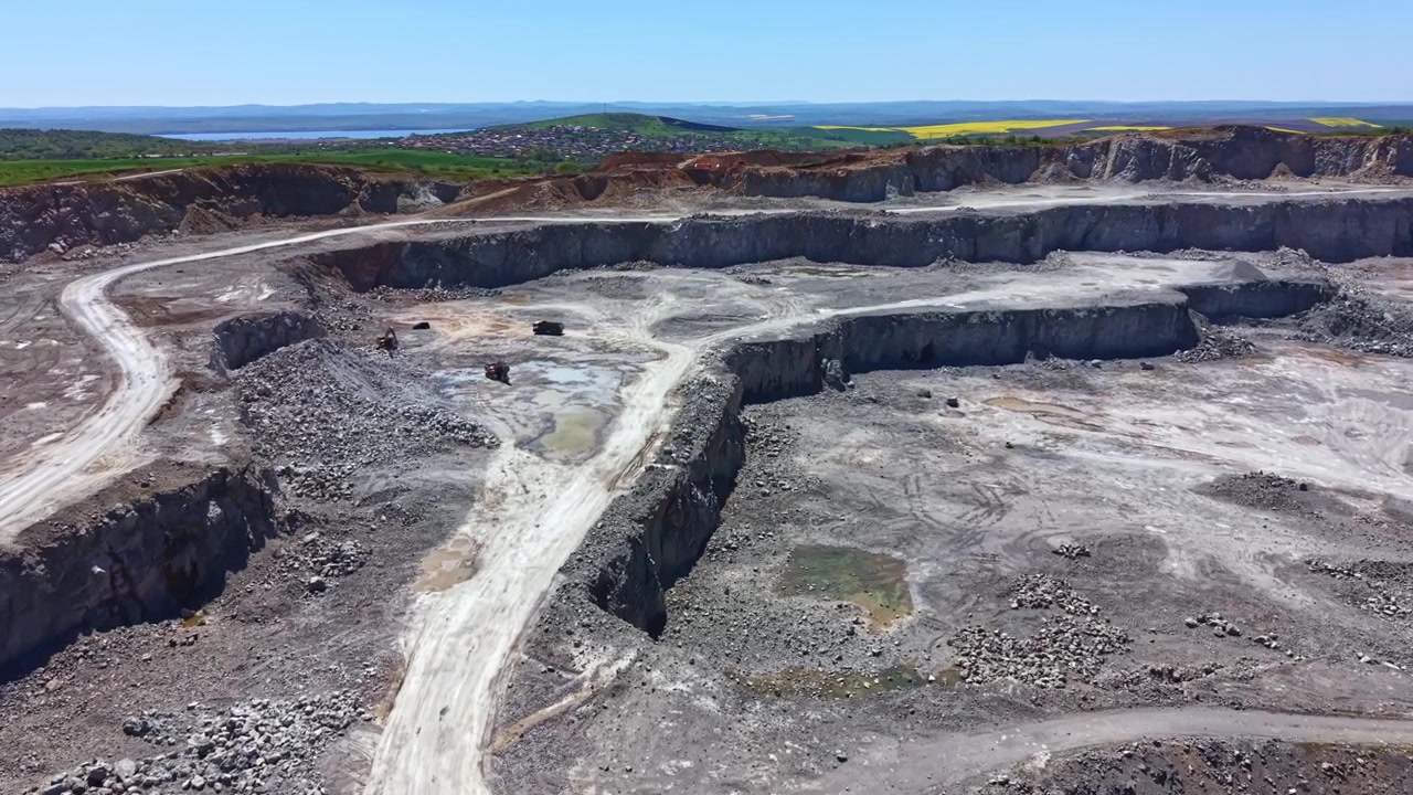 Construction work is ongoing at a large quarry site in a rural area with machines moving and dirt being displaced under a clear sky