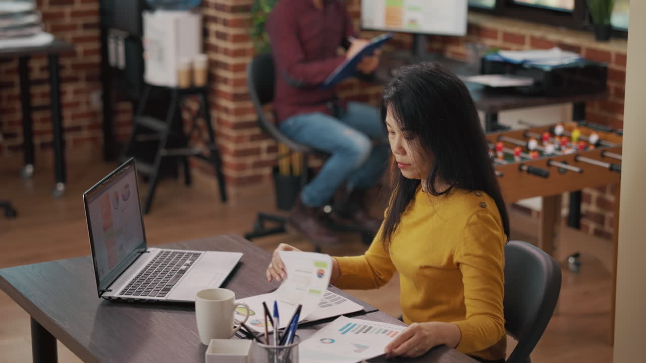 Asian woman analyzing sales charts on files and laptop