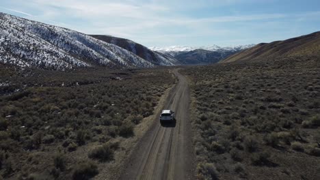 car drives down a dirt road in a desert valley in northern nevada. the dry valley floor is contrasted by snow-covered mountains, green shrubs, and sunny skies. otherworldly road trip landscape.