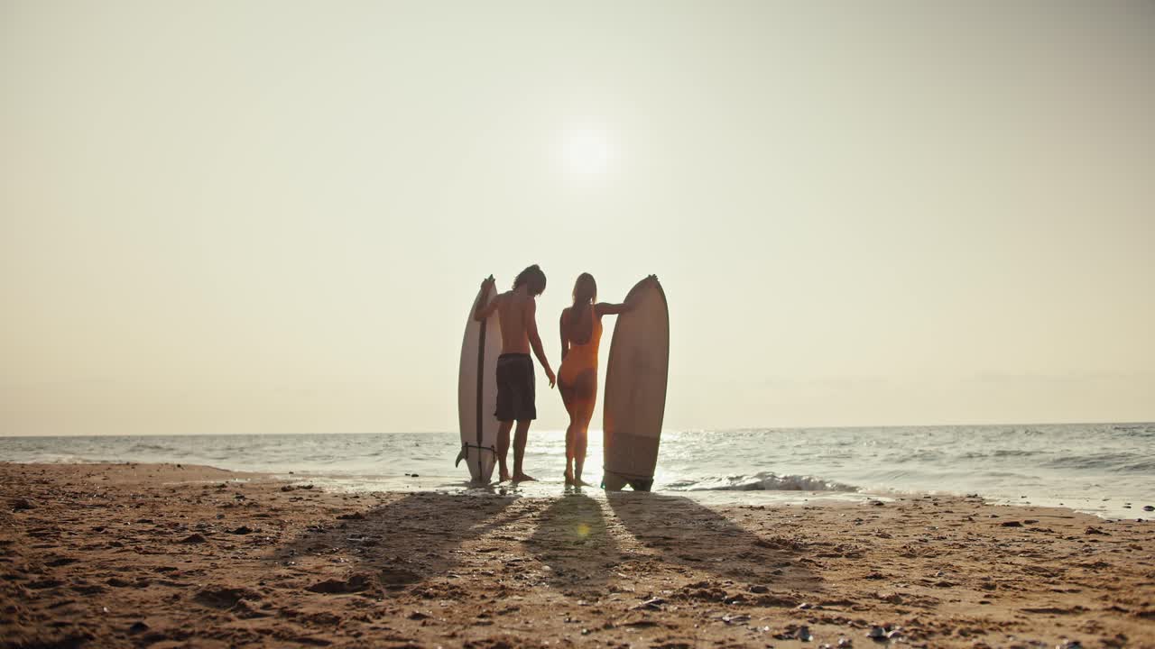 오렌지색 수영복을 입은 그의 금발 여자 친구와 함께 오렌지 모래 해변에서 바다를 향해 서서 서핑 보드를 들고 있습니다.